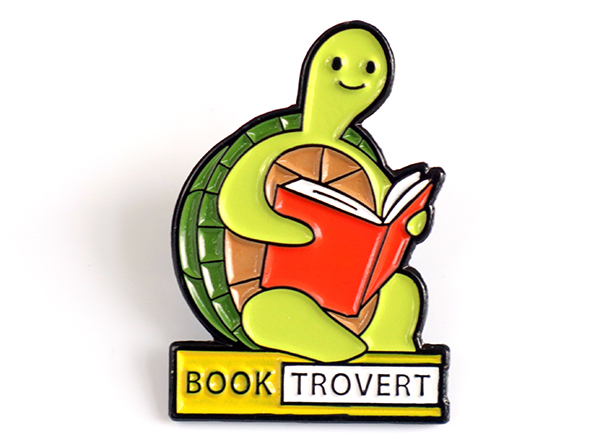 Enamel pin of a seated turtle reading a book. Beneath the turtle is the word booktrovert