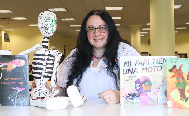 Storyteller Cris Hertle, posed with Spanish language picture books and a papier mache skeleton in the Children's area of the library.