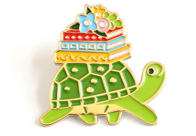 Enamel pin featuring a turtle smiling turtle carrying a stack of books and flowers on her back