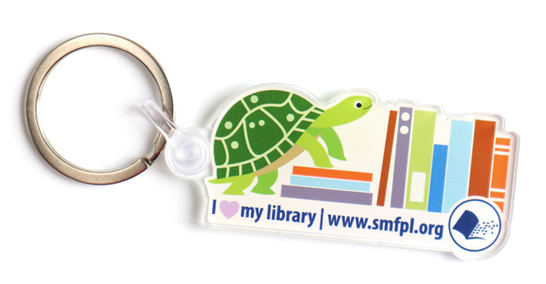 Acrylic keychain with an illustration of a smiling turtle climbing a stack of books. The text reads: I heart my library. www.smfpl.org